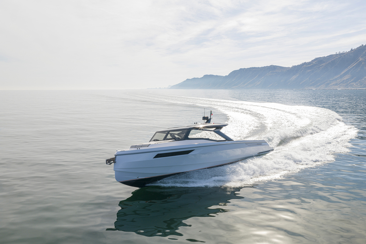 Powerful white motorboat speeding across calm coastal waters, leaving a white wake against a rocky shoreline and distant cliffs.
